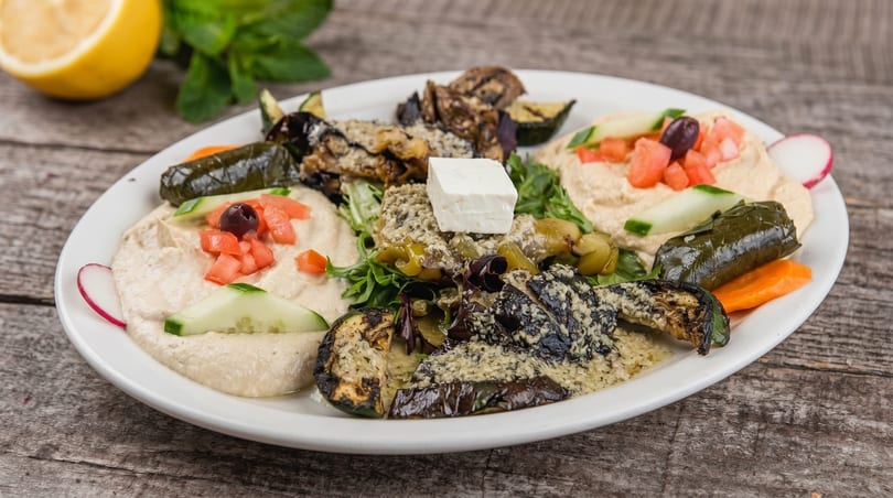 A white plate with hummus, grilled vegetables, stuffed grape leaves, and feta cheese.
