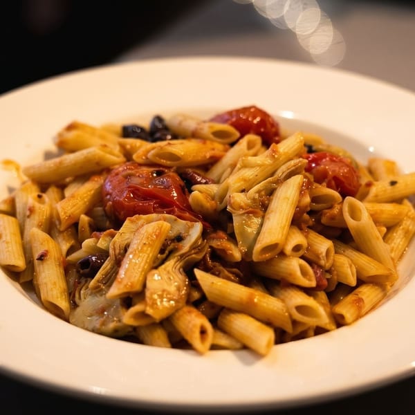 Penne pasta with roasted tomatoes, artichoke hearts, and olives in a white bowl.