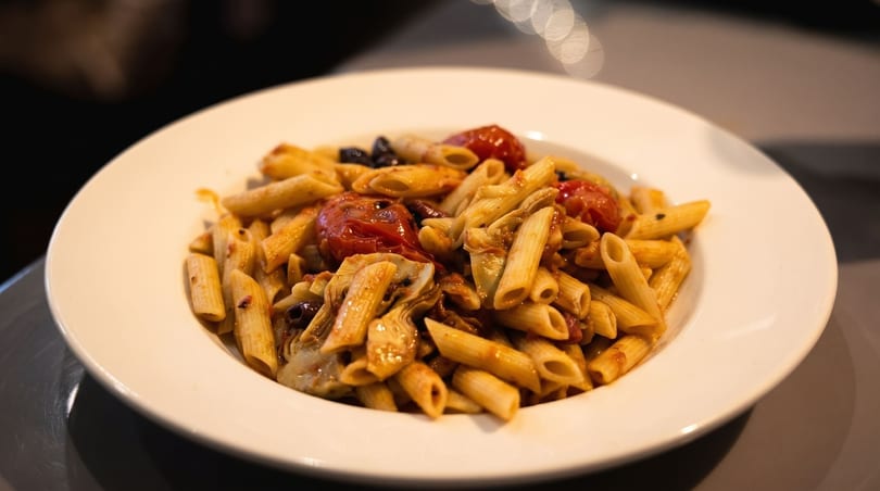 Penne pasta with roasted tomatoes, artichoke hearts, and olives in a white bowl.